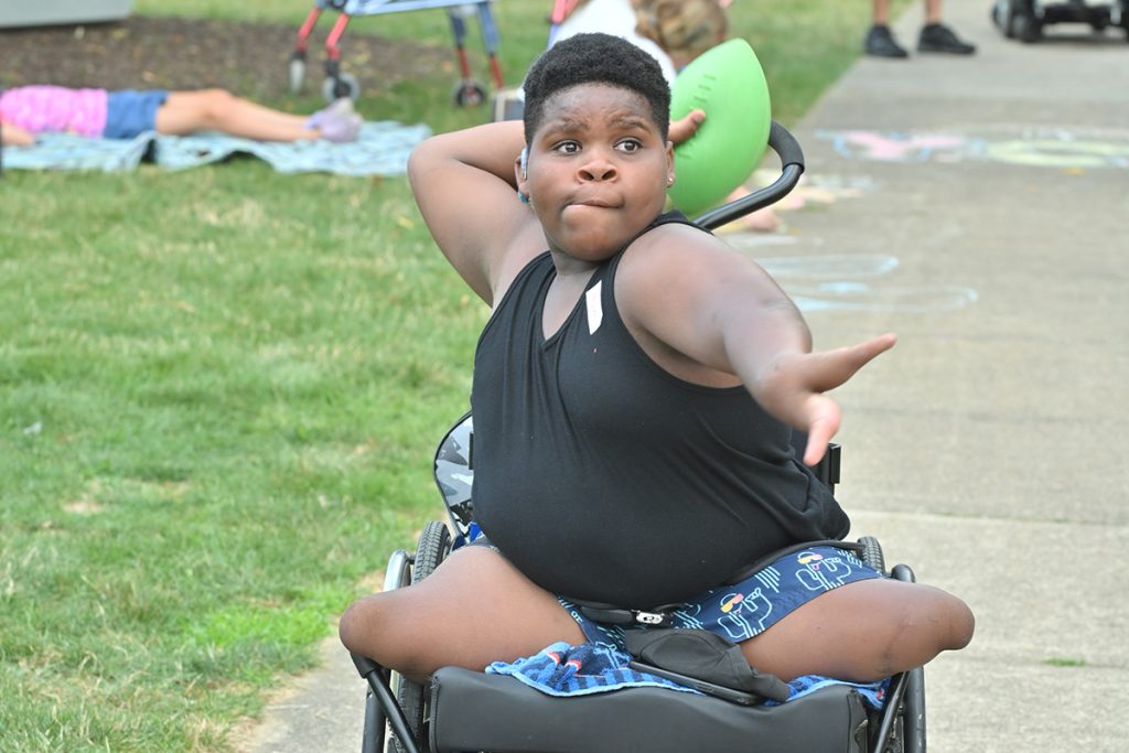 A boy in a wheelchair with a limb difference throws a football, looking focused.