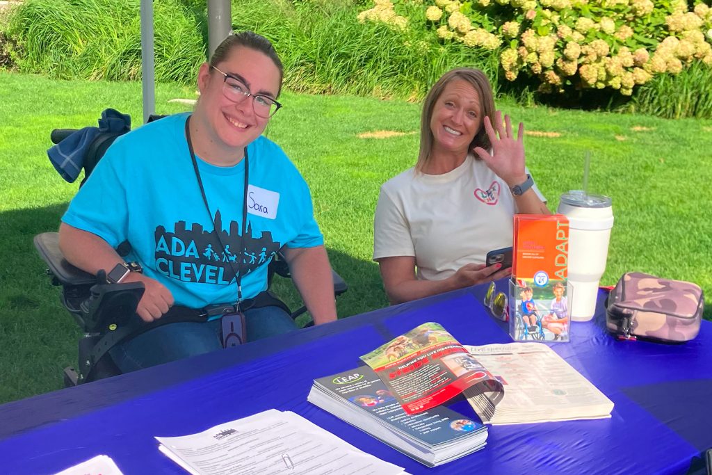 Two people sitting at a blue desk one waving to the camera