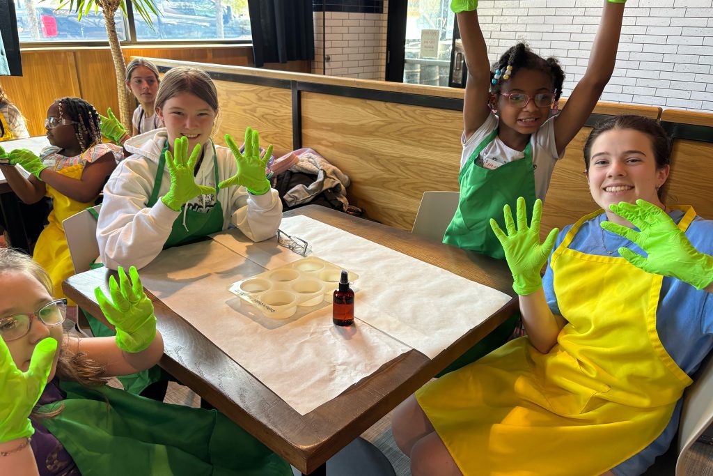 A group of kids at a table with yellow smocks on holding up their hands with green rubber gloves on