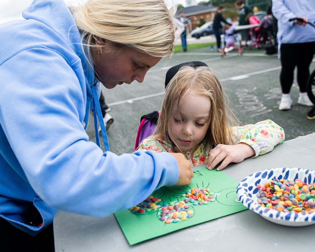 A teen leaning over helping a kid with a craft at on a green table