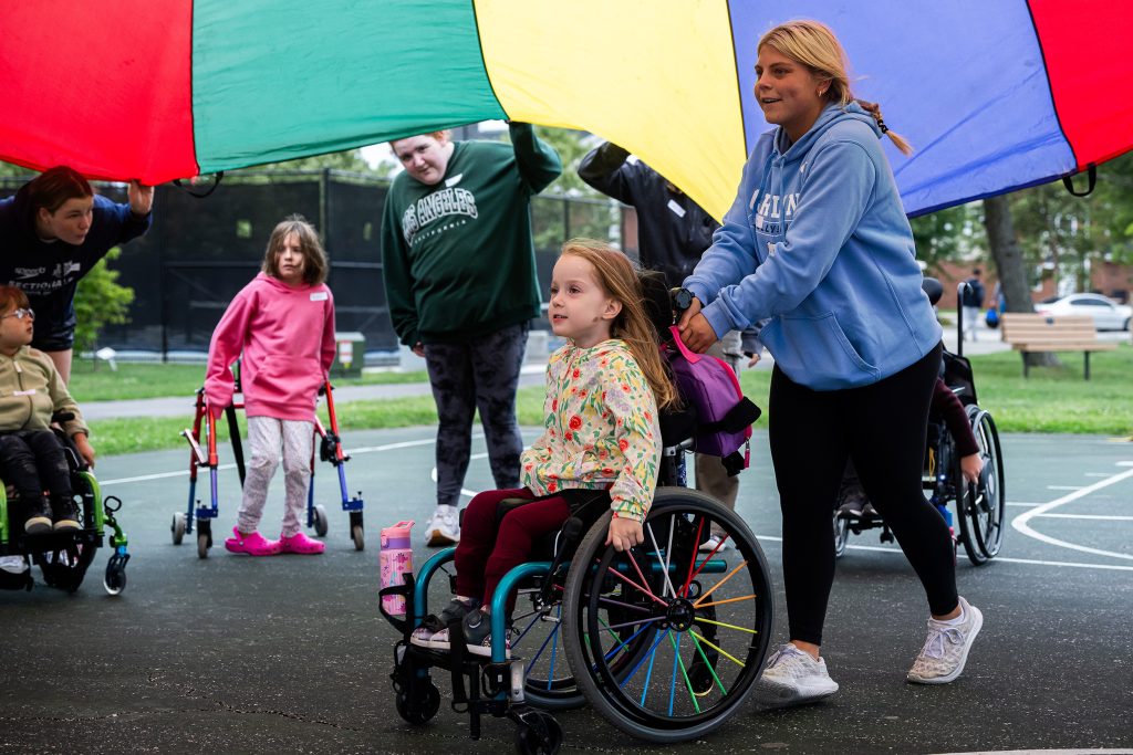 a multitude of kids and adults using a parachute under a child in a wheelchair
