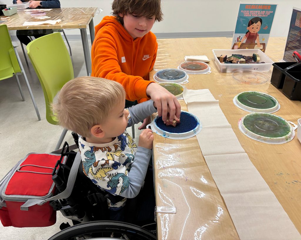 A teen in an orange hoodie sitting at a table doing crafts with a child in a wheelchair