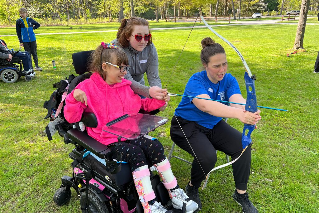Two women helping a child in a pink hoodie to use a bow and arrow