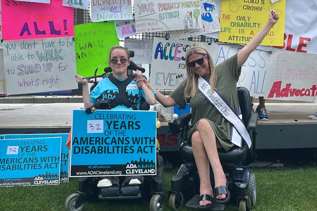Two ladies in wheelchairs holding a sign in front of a bunch of other signs