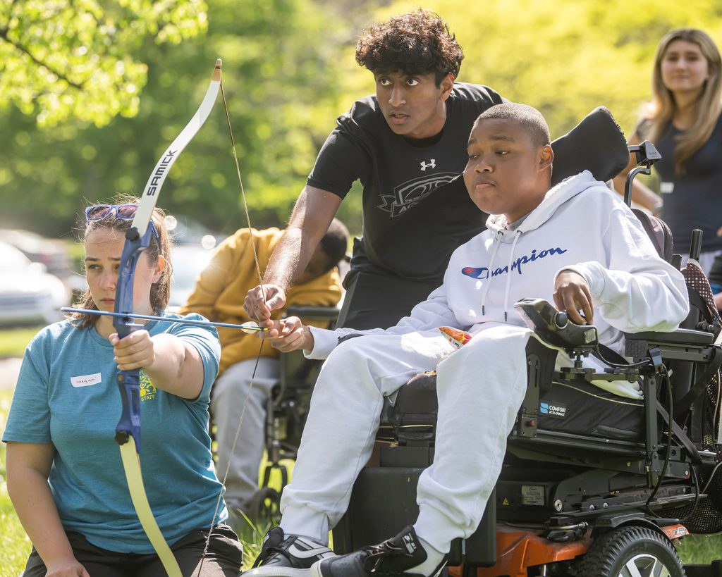 2 people helping a kid in a wheelchair with a bow and arrow