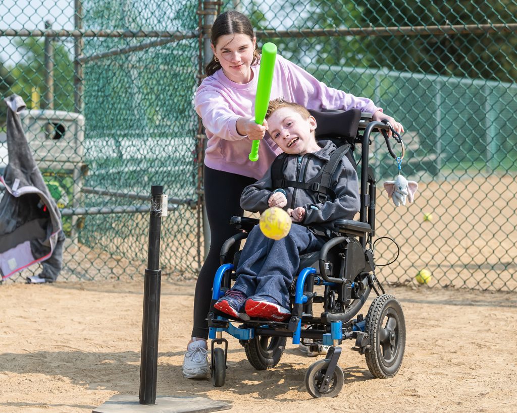 a teen helping a child in a wheelchair to play baseball