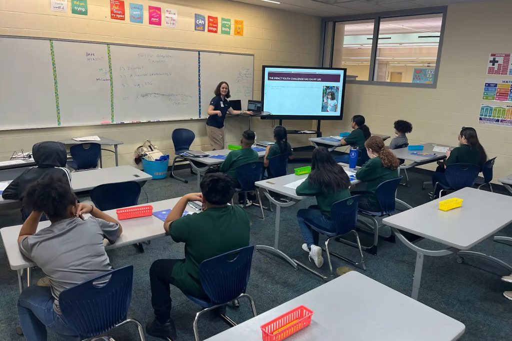 A classroom with a teacher at the front next to a TV screen
