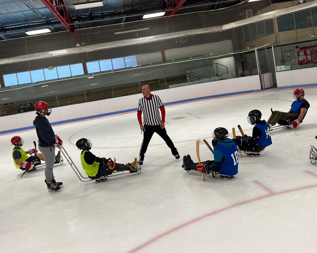 A referee flanked by kids on sleds on an ice rink playing hockey