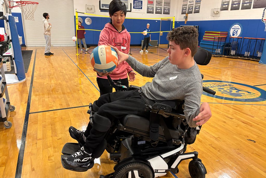 Boy in wheelchair holding a volleyball being giving by a teen boy