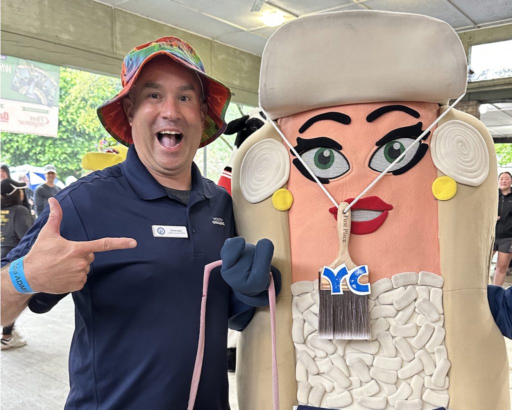 A man in a blue shirt and a hat pointing at the onion Cleveland Guardians mascot