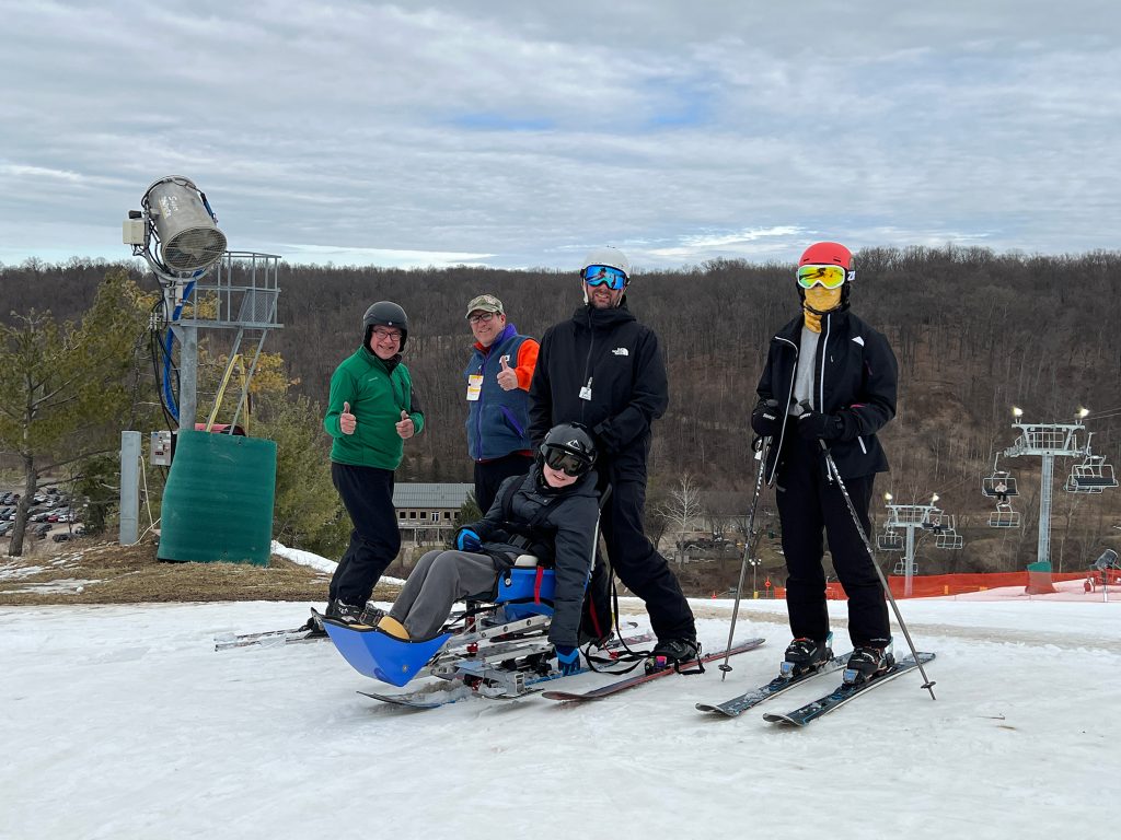 A child smiles on an accessible ski setup surrounded by four people