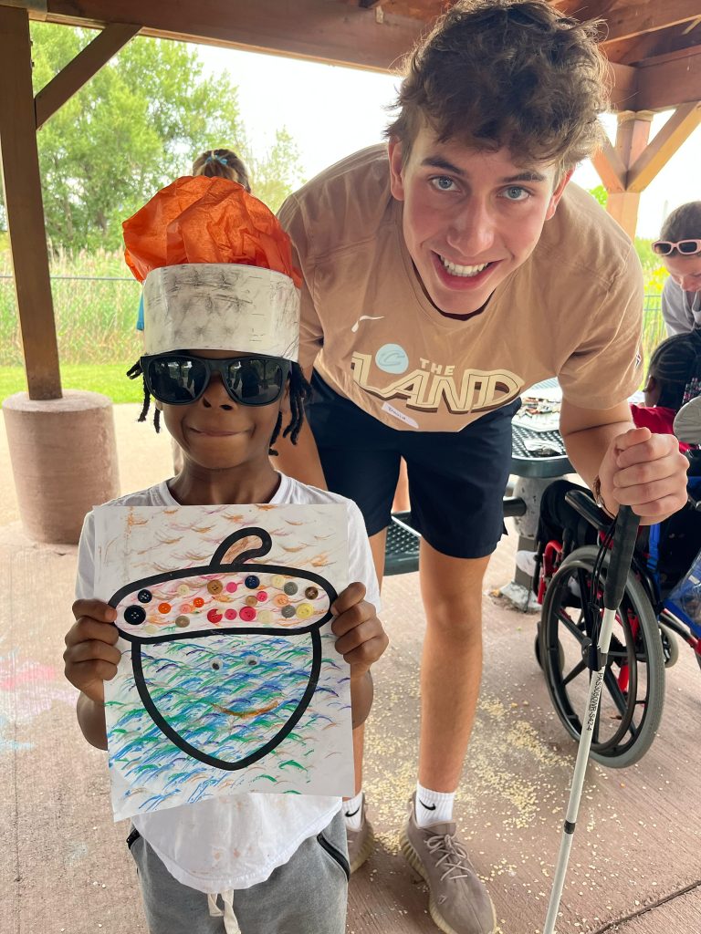 A teen smiling with a boy wearing a silly hat holding an art project