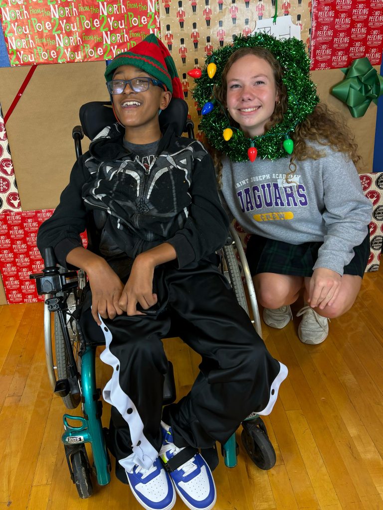 A girl with a christmas wreath around her head standing next to a boy in a wheelchair with a christmas hat on