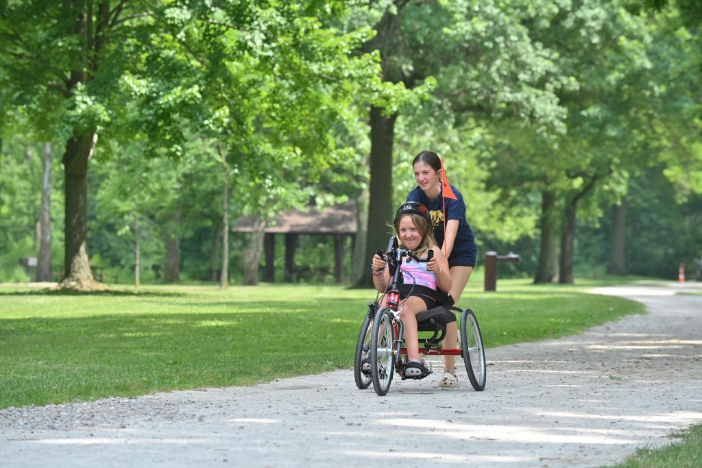A girl in a handbike with a teen pushing from behind in a park with greenery everywhere