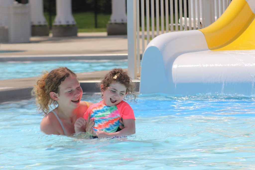 A teen holding a girl in a bright colored rash guard in a pool with a slide behind