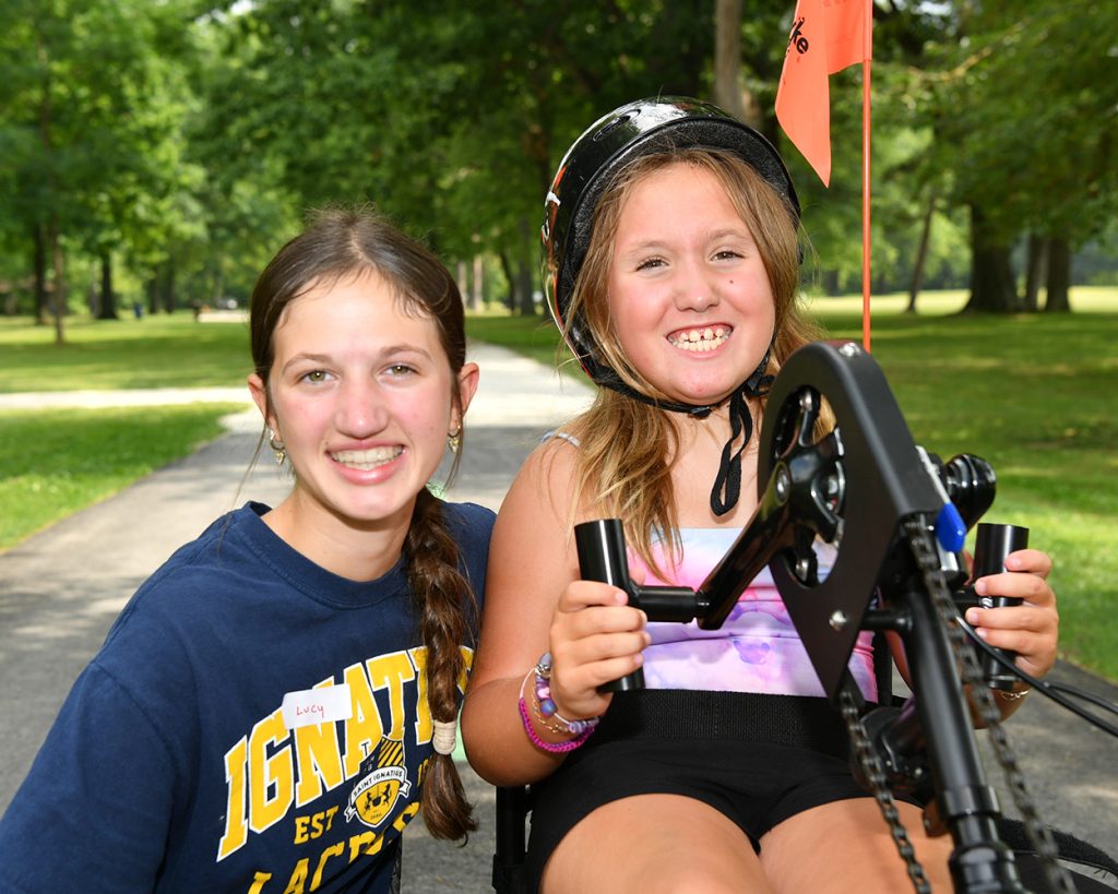 A girl on a hand bike next to a teen outside with trees in the background
