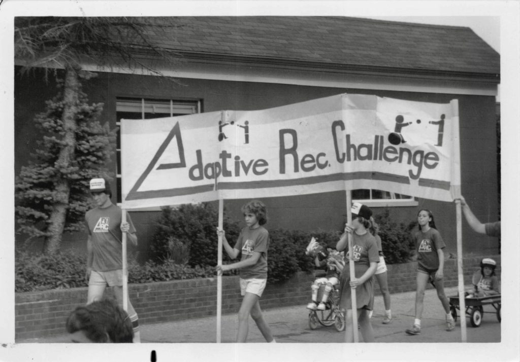 People carry a banner that says "Adapted Rec Challenge" while other people roll and walk by