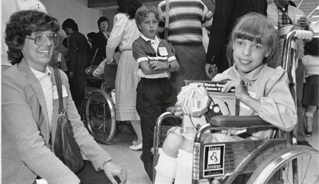 A woman sits on the floor smiling and a girl in a wheelchair smiles next to her