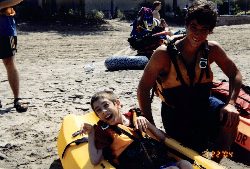 A boy sits in a kayak with a man kneeling next to him. Both are smiling and wearing life jackets