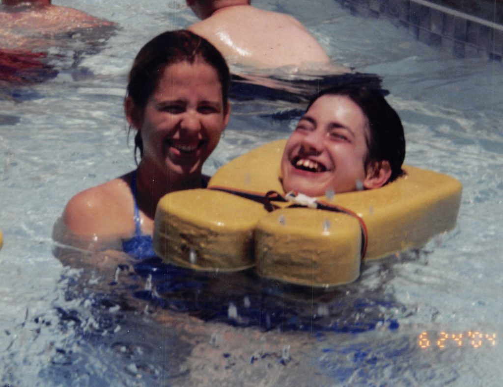 Two girls laugh in a pool. The one on the right is wearing a life jacket