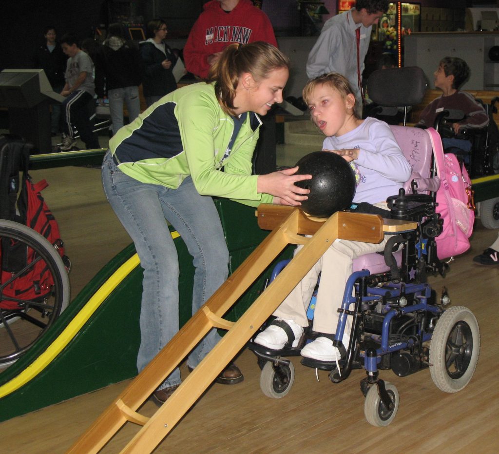 A girl leans down to help a girl in a wheelchair put a bowling ball on a ramp