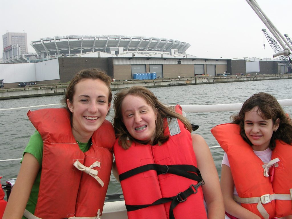 Three girls smile on a boat on the water. They are all wearing orange/red life jackets.