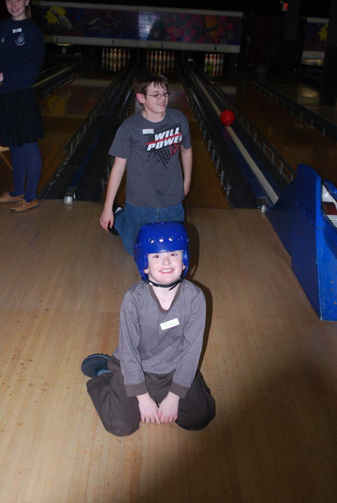 A boy stands behind another boy kneeling with a blue helmet.