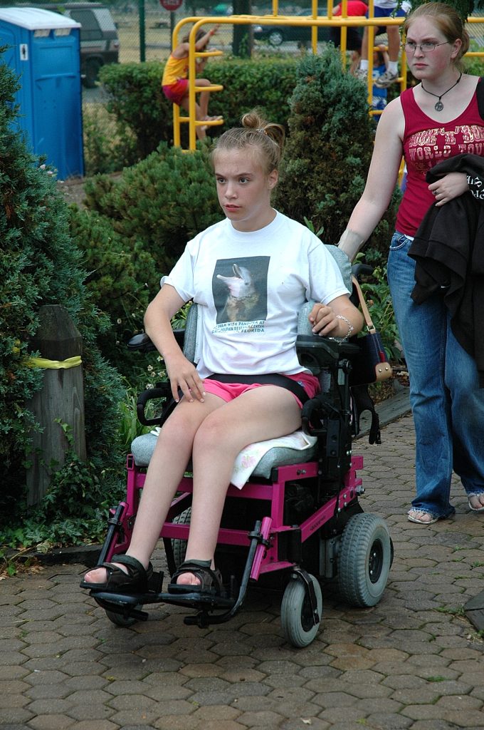 A girl in a wheelchair rolls along with a girl standing behind her.