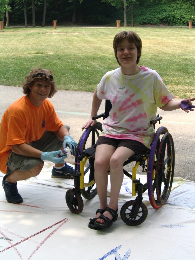 A boy in an orange shirt crouches on a piece of paper next to a girl in a wheelchair with paint on her hand.