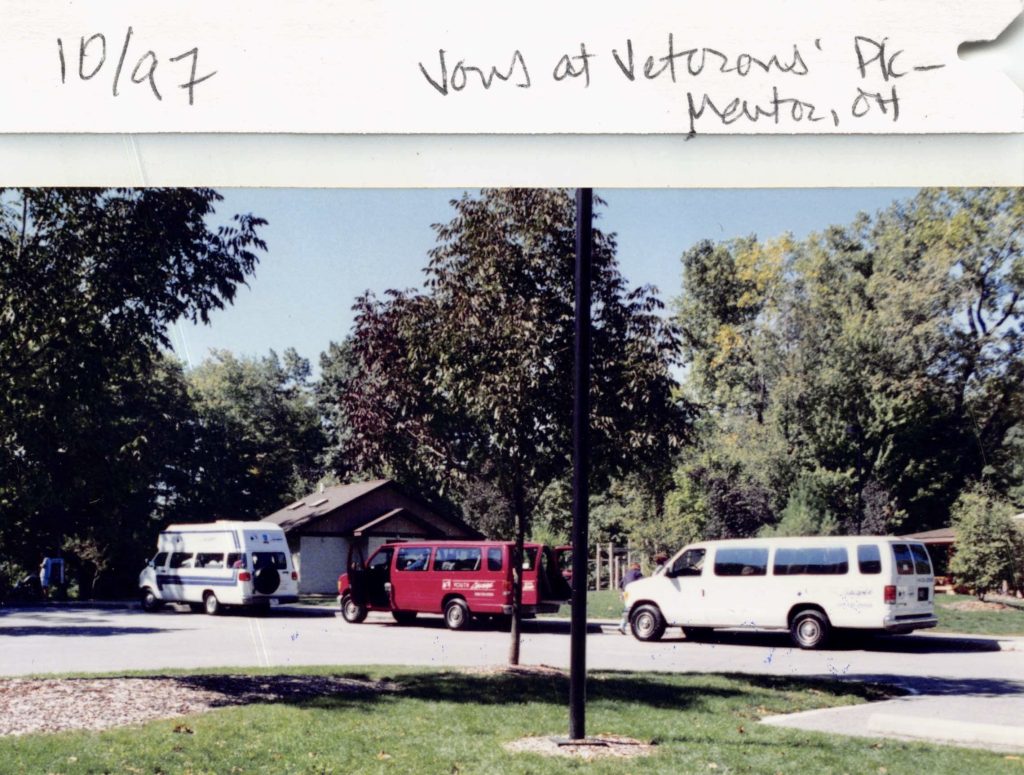 Three vans - white, red, and white, linked up on a street near some grass and trees