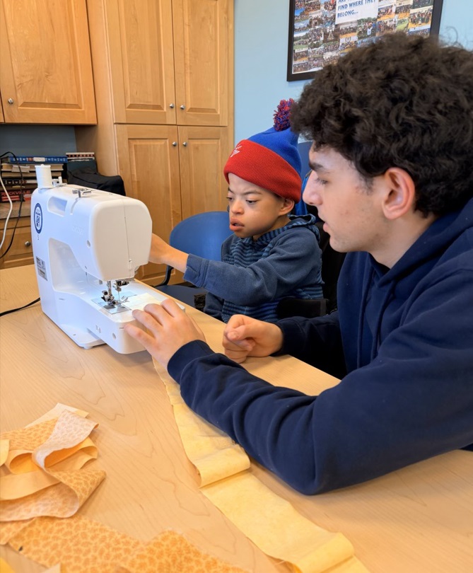 A YC participant presses a button on a sewing machine as his volunteer prepares to thread a yellow cloth through
