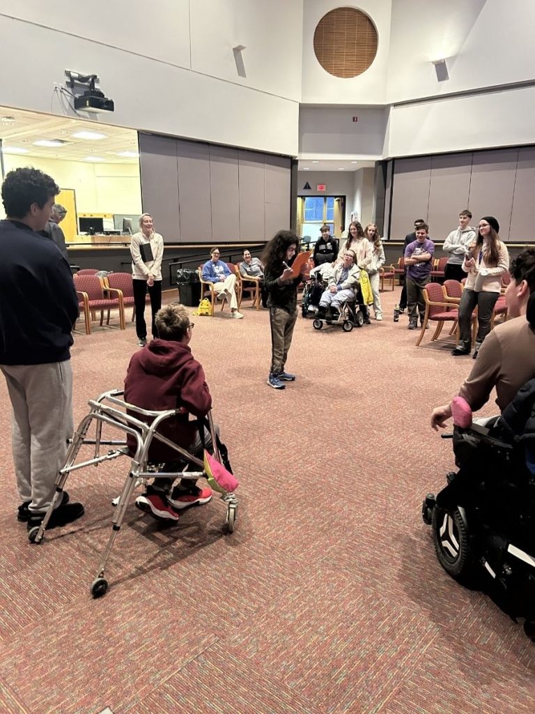 A teen boy with long hair looks intently at a clipboard surrounded by a circle of YC participants and volunteers