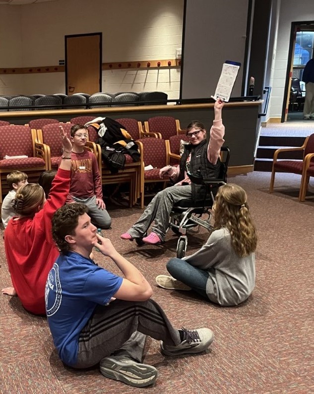 A participant in a wheelchair holds a clipboard in the air, while others sit around her.