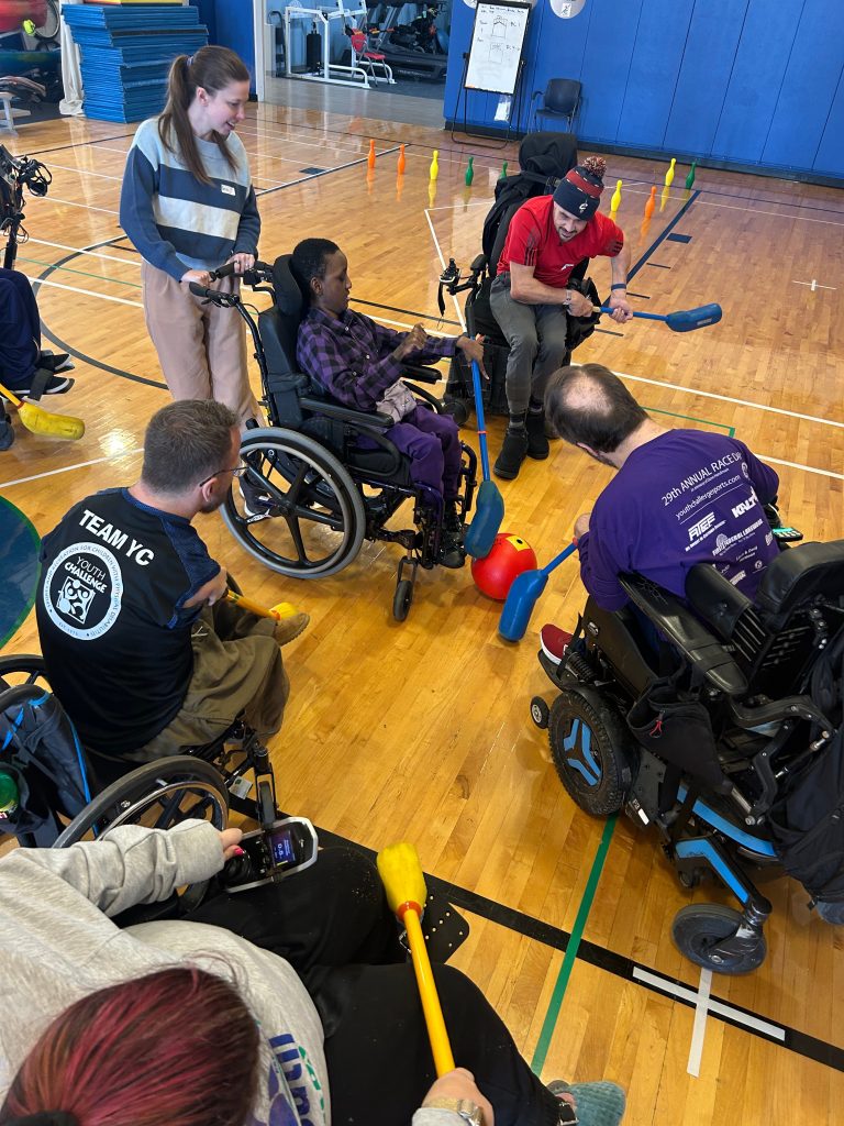 A group of adults play gym hockey in the YC gym. Most people are using wheelchairs and attempting to move a red ball with blue foam hockey sticks.