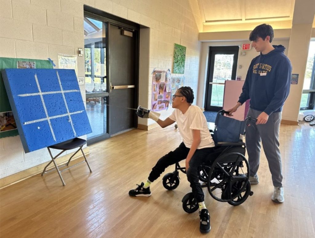young man in wheelchair holds out a foil (sword) to a target as a volunteer stands behind
