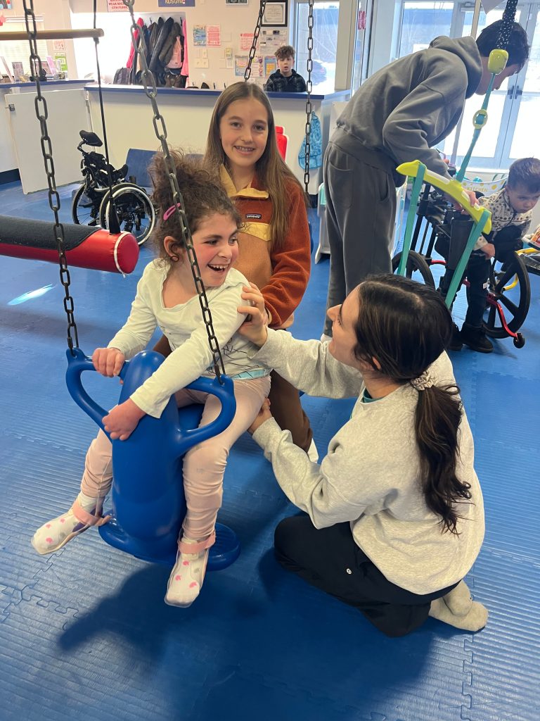 Two girls sit on a blue swing smiling while another girl sits on the floor.
