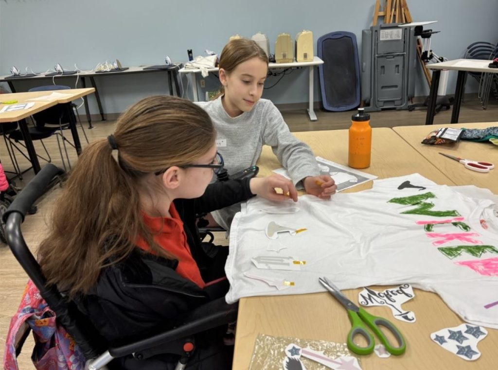 two young women decorate a shirt with "Wicked" written in marker, one of the women is a wheelchair user