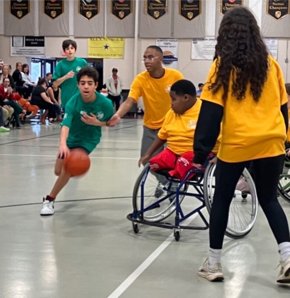 Immanuel dribbles a basketball past yellow-team defenders Marcus, Brycen, and Kylie during a game in a crowded gymnasium.