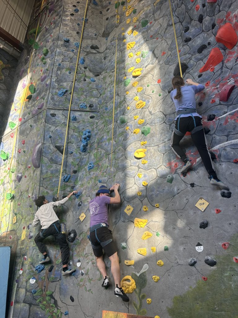 Immanuel and two other people climb a rock climbing wall.