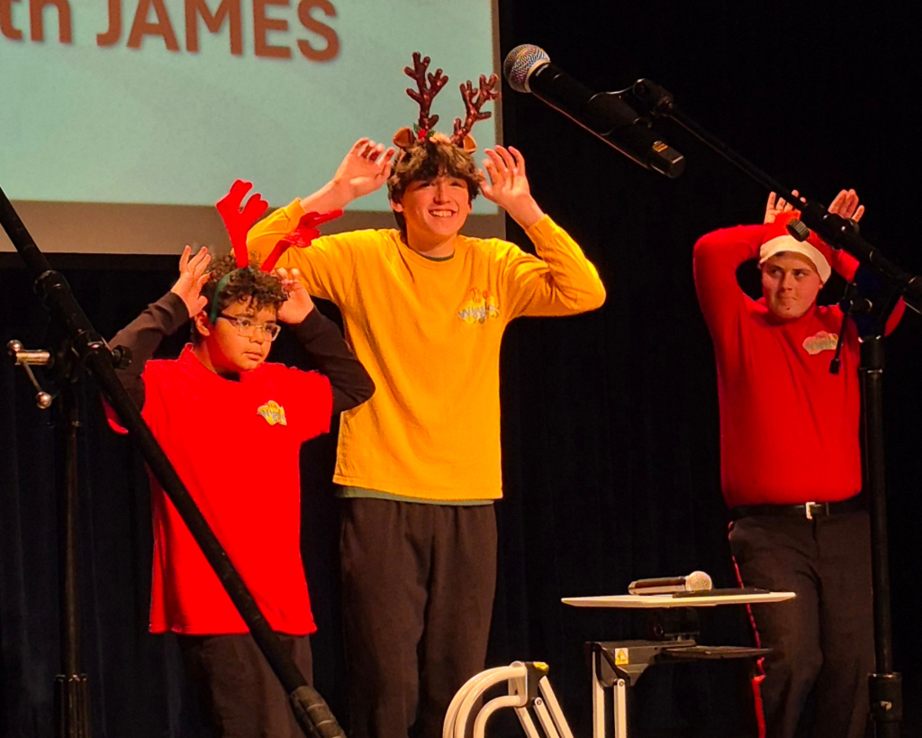 Three boys stand on a stage with their hands on their heads