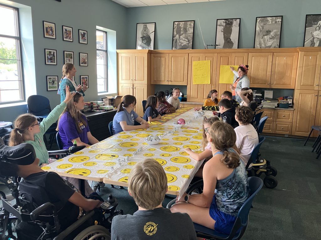A group of participants and volunteers sit at table in volunteer room as YC staffer Megan stands in front. There is a smiley face table cloth on the table, 70s theme,