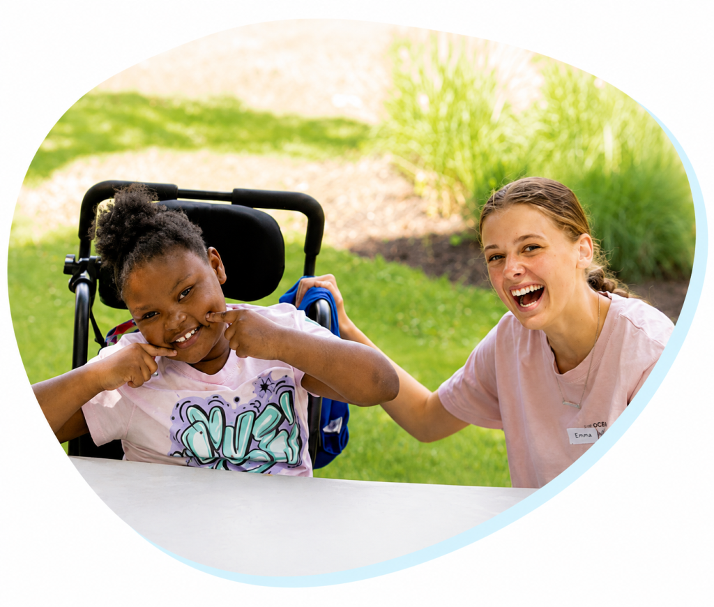 A girl in a wheelchair points to her cheeks and smiles while another girl sits next to her smiling.